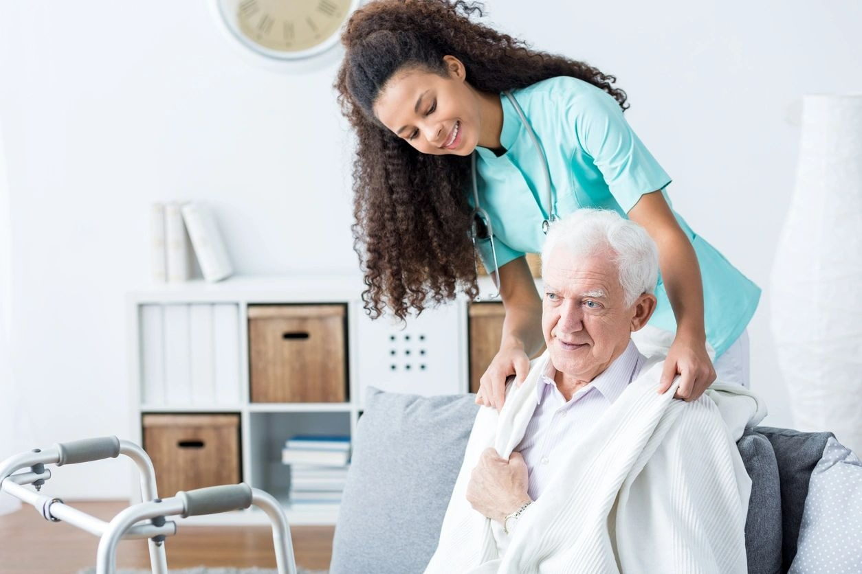 A caregiver gently supports an elderly man in a cozy living room.