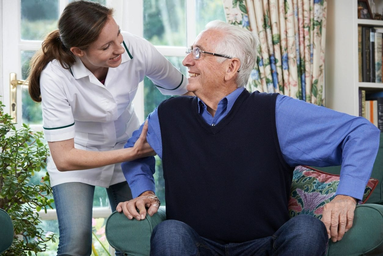 A nurse smiling and assisting an elderly man sitting on a chair.